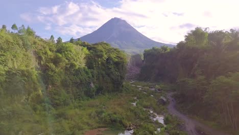 Tiro-De-Dron-Ascendente-Sobre-Un-Lago-Natural-En-La-Naturaleza-Con-Volcán-Merapi-Y-Camino-De-Lava-Y-Carretera-Durante-El-Verano