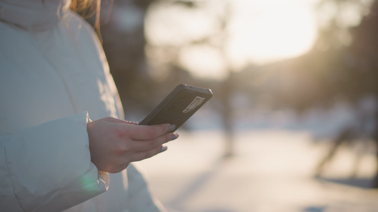 Close up shot of woman wearing winter coat and long manicured nails pressing phone outdoors against blurred snowy background with sunlight glow evoking calm joyful mood