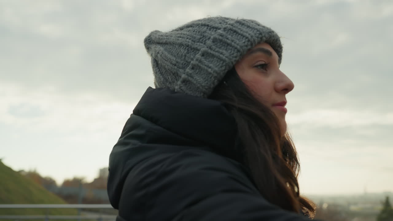 Closeup portrait of woman on bridge smiling with beanie and jacket, soft gaze toward skyline, warm expression and subtle backlight, candid urban moment with bench and lamp in frame