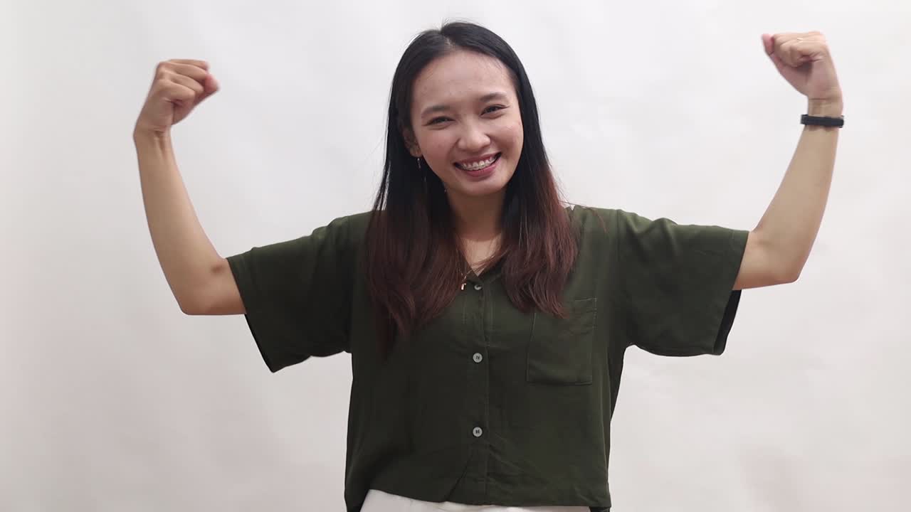 Strong young woman of Asian ethnicity wear green shirt showing biceps muscles on hand demonstrating strength power isolated on white background studio portrait