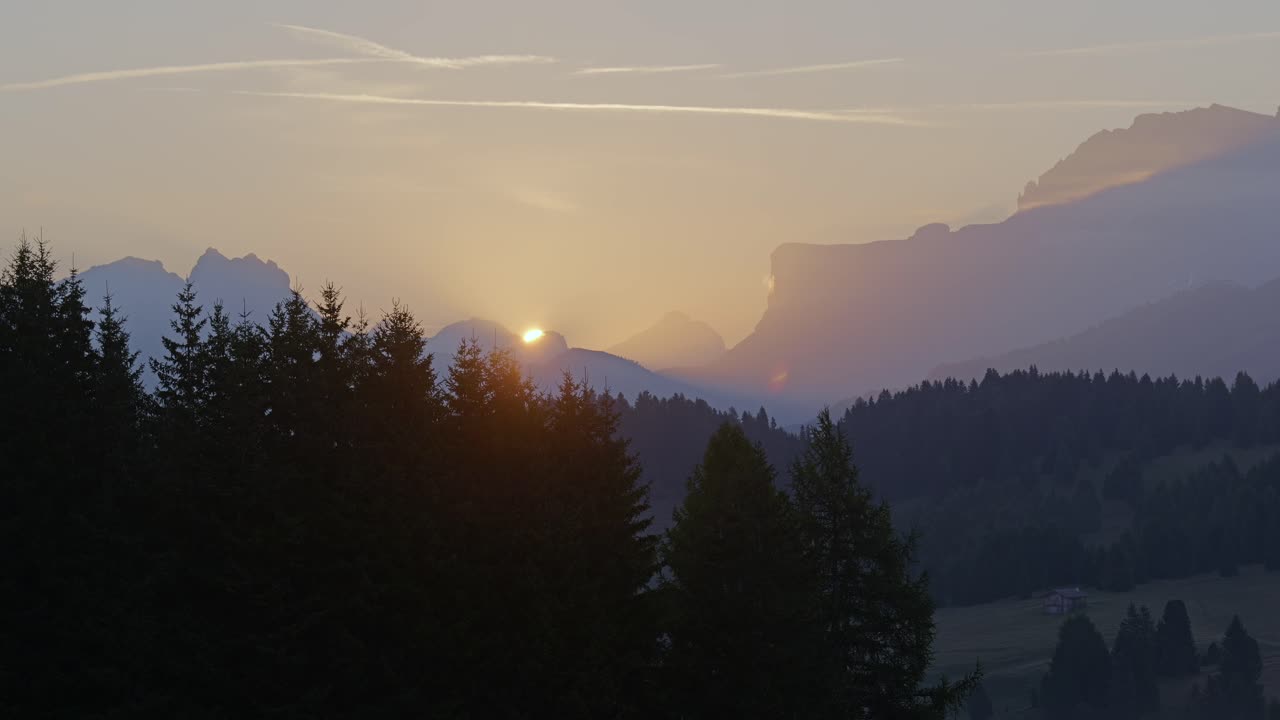 Sun rises over Seiser Alm in the Dolomites, casting warm light over misty peaks and silhouetted pine trees. This peaceful morning scene captures the beauty of Italy’s alpine landscape.