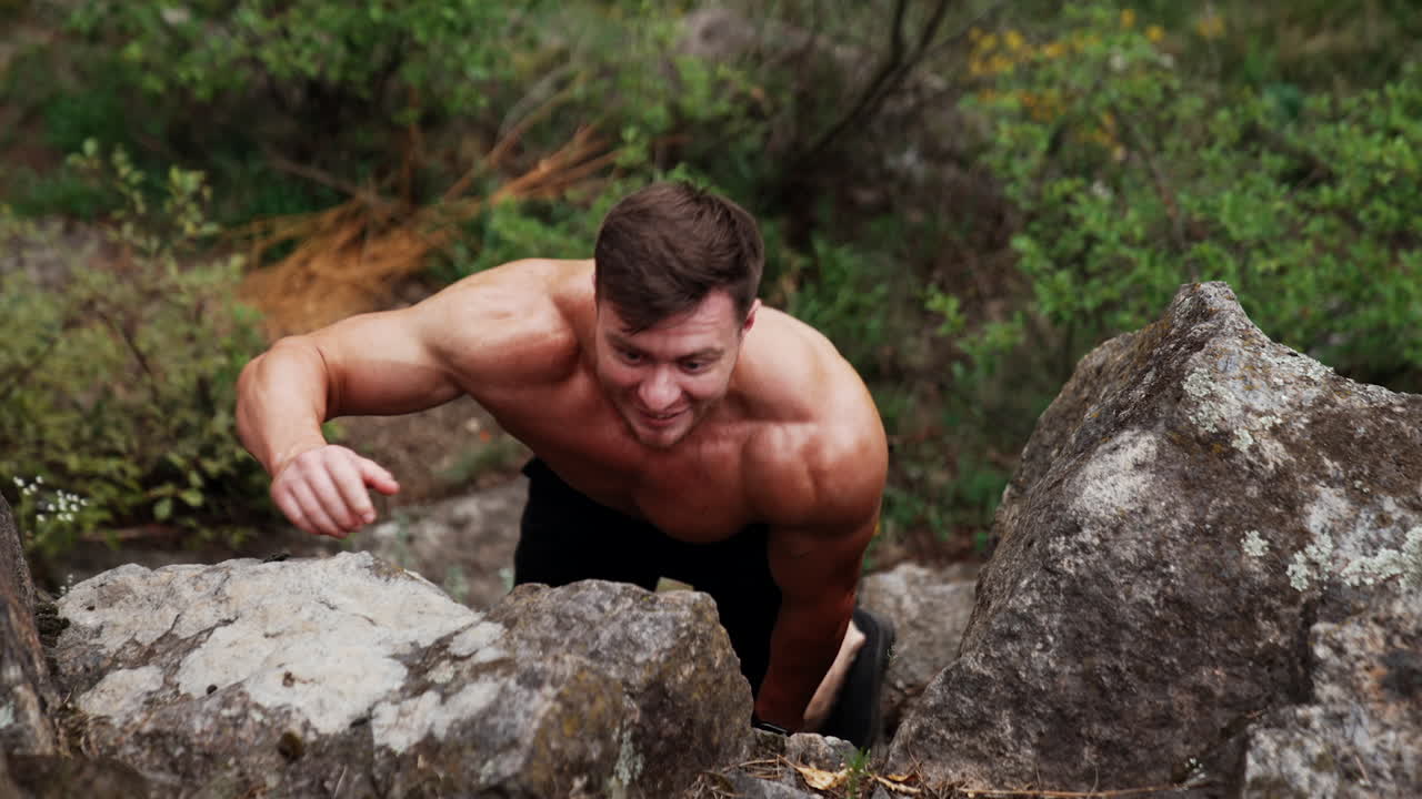 Sportive Caucasian man with naked torso climbs the high rock. Strongman uses nature surrounding for work out.