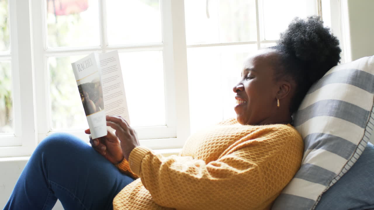 Happy african american senior woman lying, reading book and smiling in sunny room, slow motion