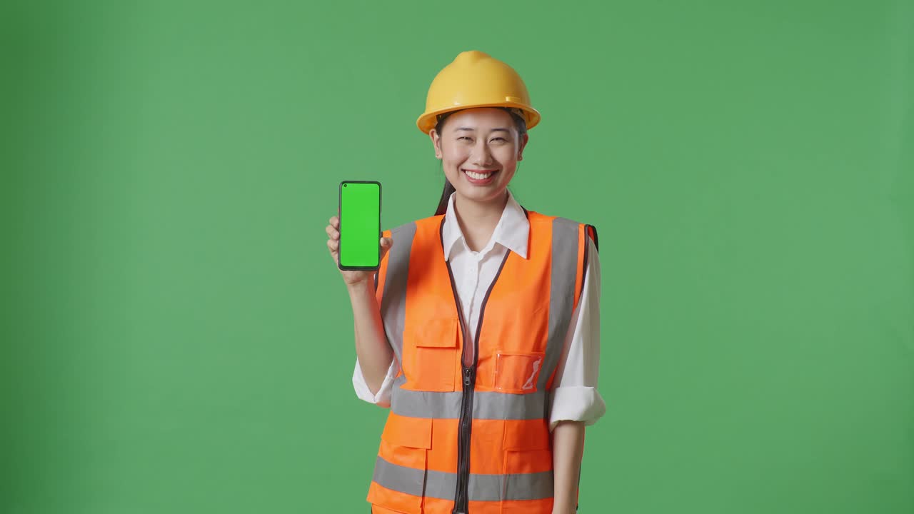 Asian Female Engineer With Safety Helmet Smiling And Showing Green Screen Smartphone To The Camera While Standing In The Green Screen Background Studio