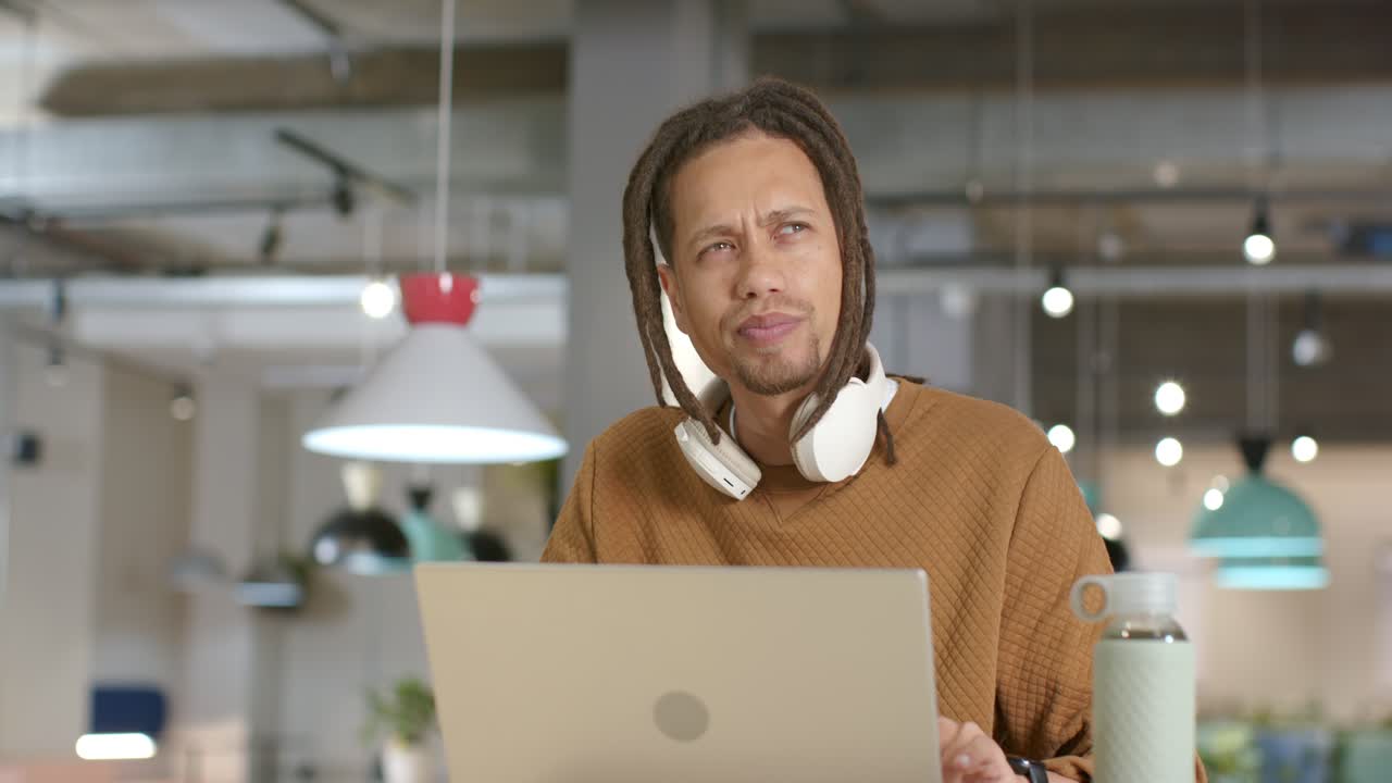 African American man thinking at office desk, adjusting headphones and composing on laptop for work