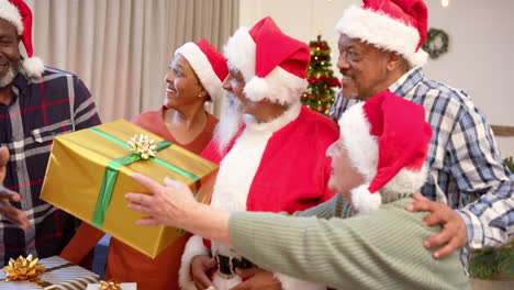 Happy-diverse-senior-friends-in-santa-outfit-and-christmas-hats-exchanging-gift-at-home,-slow-motion