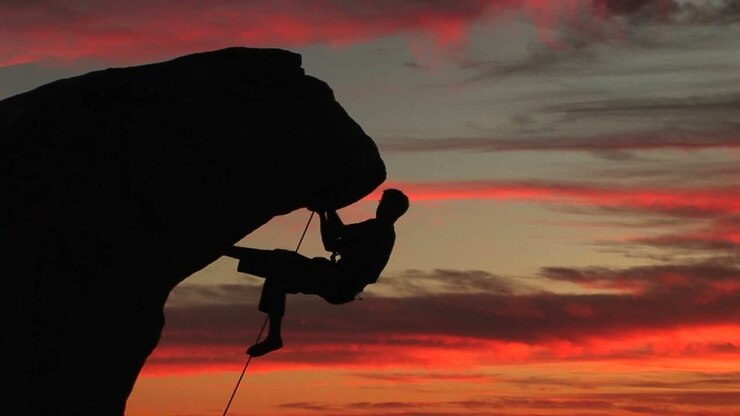 Inquadratura media di un alpinista in controluce appeso a una parete rocciosa a strapiombo con un magnifico tramonto californiano in lontananza.