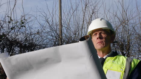 Close-up-portrait-of-an-mature-senior-architect-building-inspector-inspecting-a-construction-site-with-architectural-plans-and-hard-hat