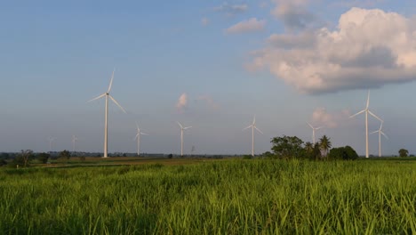 Windkraftanlagen-Schießen-Aus-Einem-Ackerland,-Während-Die-Sonne-Untergeht,-Während-Lastwagen-Vorbeifahren-Und-Staubwolken-Erzeugen,-Saubere-Alternative-Energie-In-Thailand-Und-Auf-Dem-Südostasiatischen-Festland