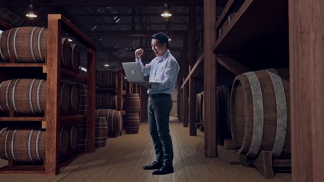 full body side view of an asian male professional worker standing with his laptop in cellar of winery, he raises his fist up with screaming goal after typed on the laptop