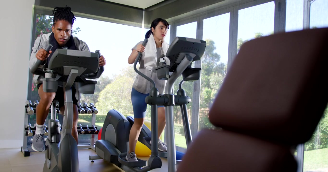 Compagni di palestra che pedalano su una cyclette e usano un'ellittica in palestra dopo essere saliti, facendo cardio con un asciugamano.