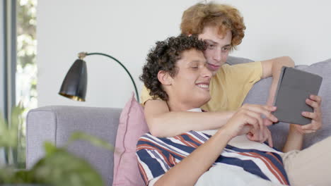 happy diverse gay male couple sitting at sofa using tablet at home, slow motion