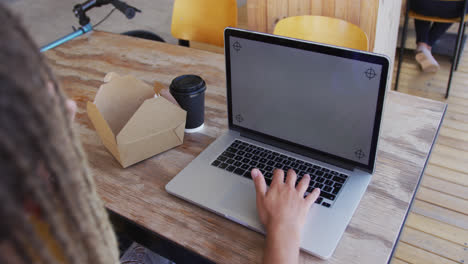 mixed race man with dreadlocks sitting in cafe with sandwich and laptop with copy space on screen