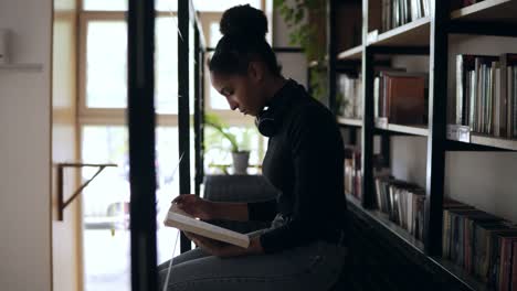 Estudiante-Elegante-Recopilando-Información-De-Diferentes-Libros,-Chica-Elegante-Con-Suéter-Negro-Y-Auriculares-Alrededor-Del-Cuello-Recopilando-Datos-Para-Los-Libros,-Sentada-En-El-Nivel-Superior-Interior-En-Una-Biblioteca-Moderna