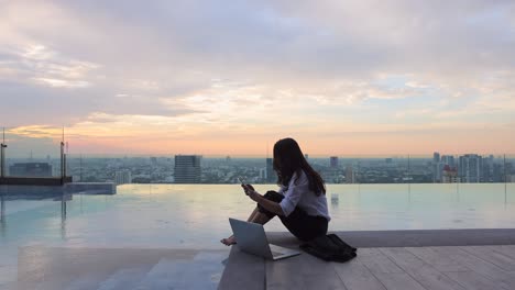 summer day lifestyle asian woman relax and chill near luxury pool on skyscraper. asian woman using app on mobile phone and working on a laptop and blurred background skyscraper, beautiful sky.