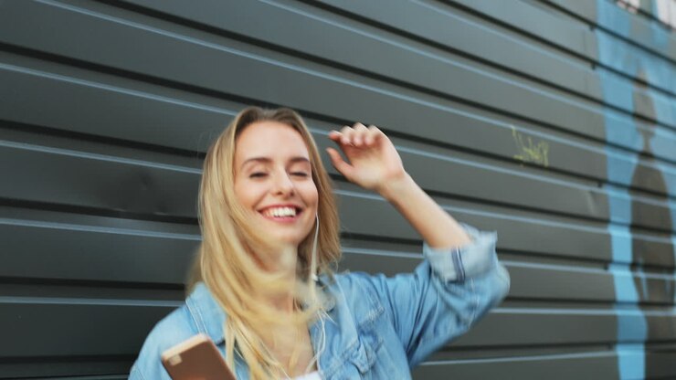 Giovane donna bionda che ascolta musica dallo smartphone e balla per strada.