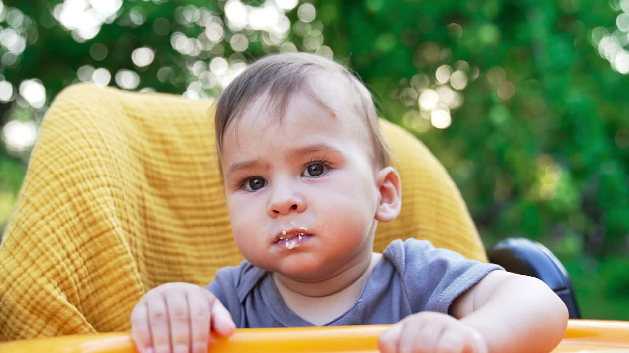 Mom is feeding her cute son with some dairy products from spoon. Lovely boy in yellow chair eating and looking intently into camera. Close up.