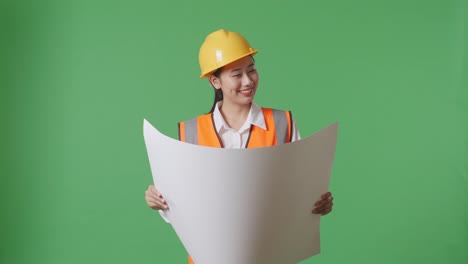 asian female engineer with safety helmet looking at blueprint in her hands and looking around while standing in the green screen background studio