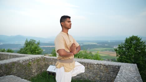 concentrated-open-eyed-Indian-man-doing-a-hatha-yoga-pose-standing-on-one-leg-overlooking-the-valley-from-a-top-of-a-hill-at-sunrise-at-dawn