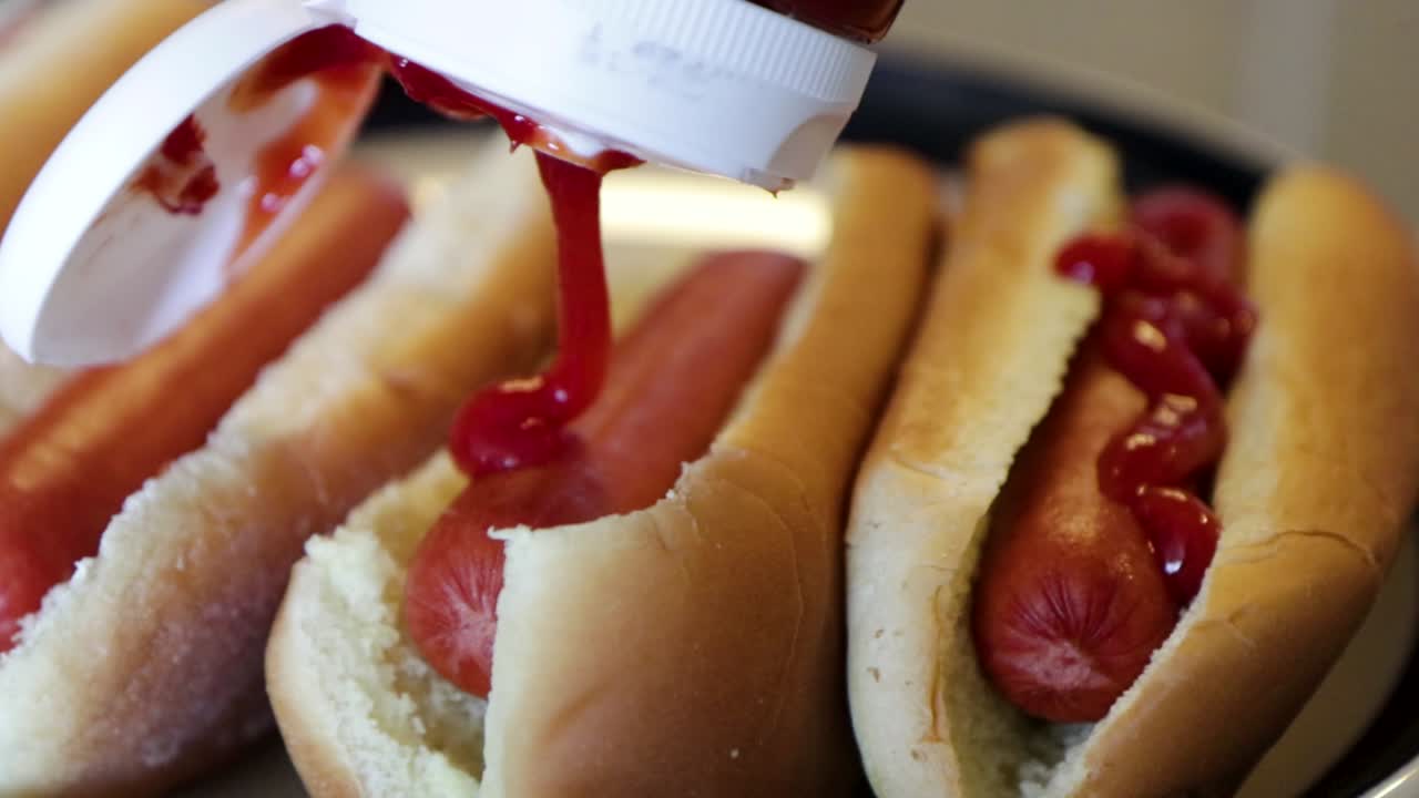 Close-up view pouring tomato ketchup over fresh homemade hotdogs in a swirly motion.