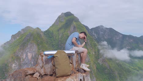 sad asian hiker male with a backpack sitting on the rock of top foggy mountain