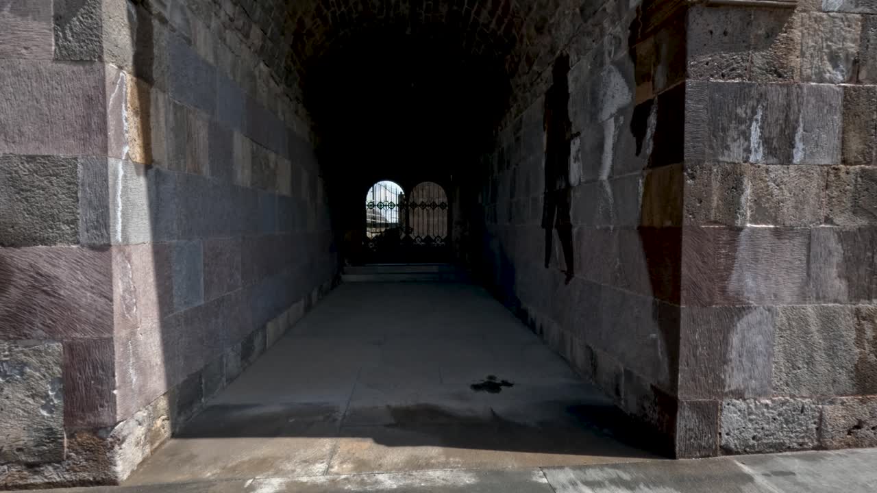 Uma pessoa caminha por um antigo arco de pedra com vista para o exterior. A luz do sol entra, mostrando a textura das pedras e o caminho. Os arredores incluem grama e algumas estruturas