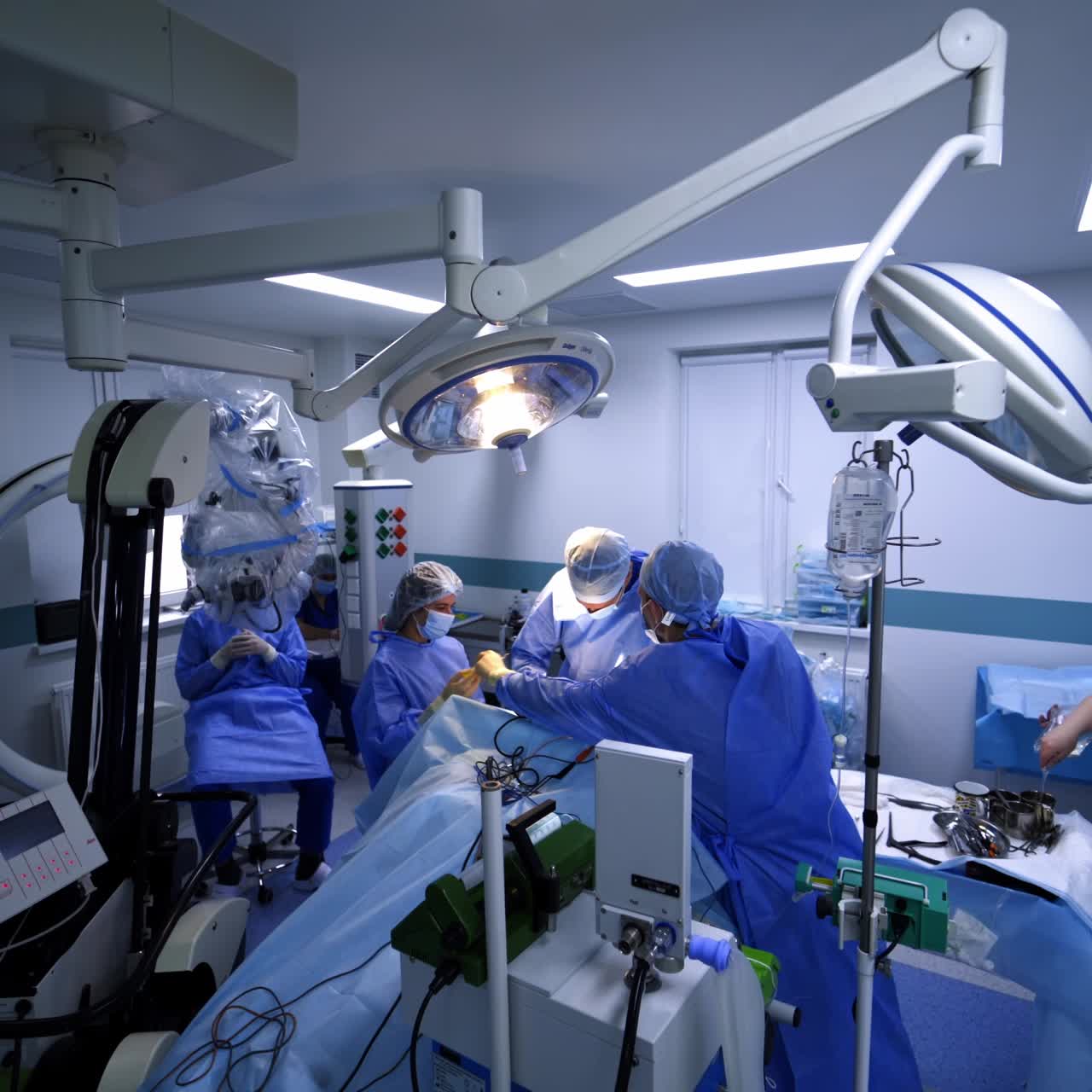 Three doctors stand over the patient on operational table. Assistants sit at backdrop in modern highly equipped surgery room