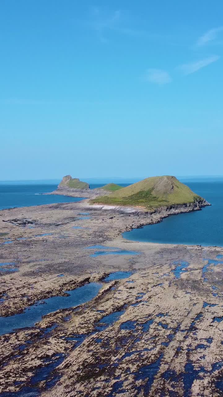 Vertical Video of Worm's Head in Rhossili at Low Tide with Grassy Hill Cut Off as Water Rises. Beautiful Blue Sky Holiday Tourism Footage.
