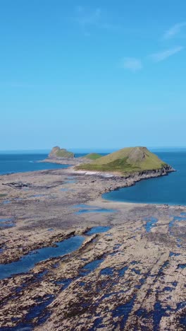 vertical video of worm's head in rhossili at low tide with grassy hill cut off as water rises. beautiful blue sky holiday tourism footage.