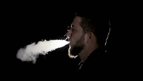 un joven barbudo blanco con camisa negra está vapeando un cigarrillo electrónico
