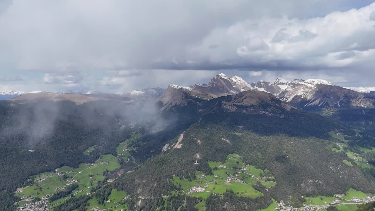 Alpe di Siusi drone footage shows Ortisei and Seceda in the Dolomites, with foggy valleys, green meadows, and snow-capped peaks under a cloudy sky