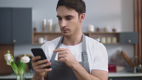 Retrato-De-Un-Chef-Sonriente-Mirando-Una-Receta-En-Un-Teléfono-Móvil-En-La-Cocina-De-Casa.