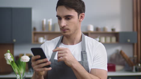 retrato de un chef sonriente que busca una receta en un teléfono móvil en la cocina de su casa.