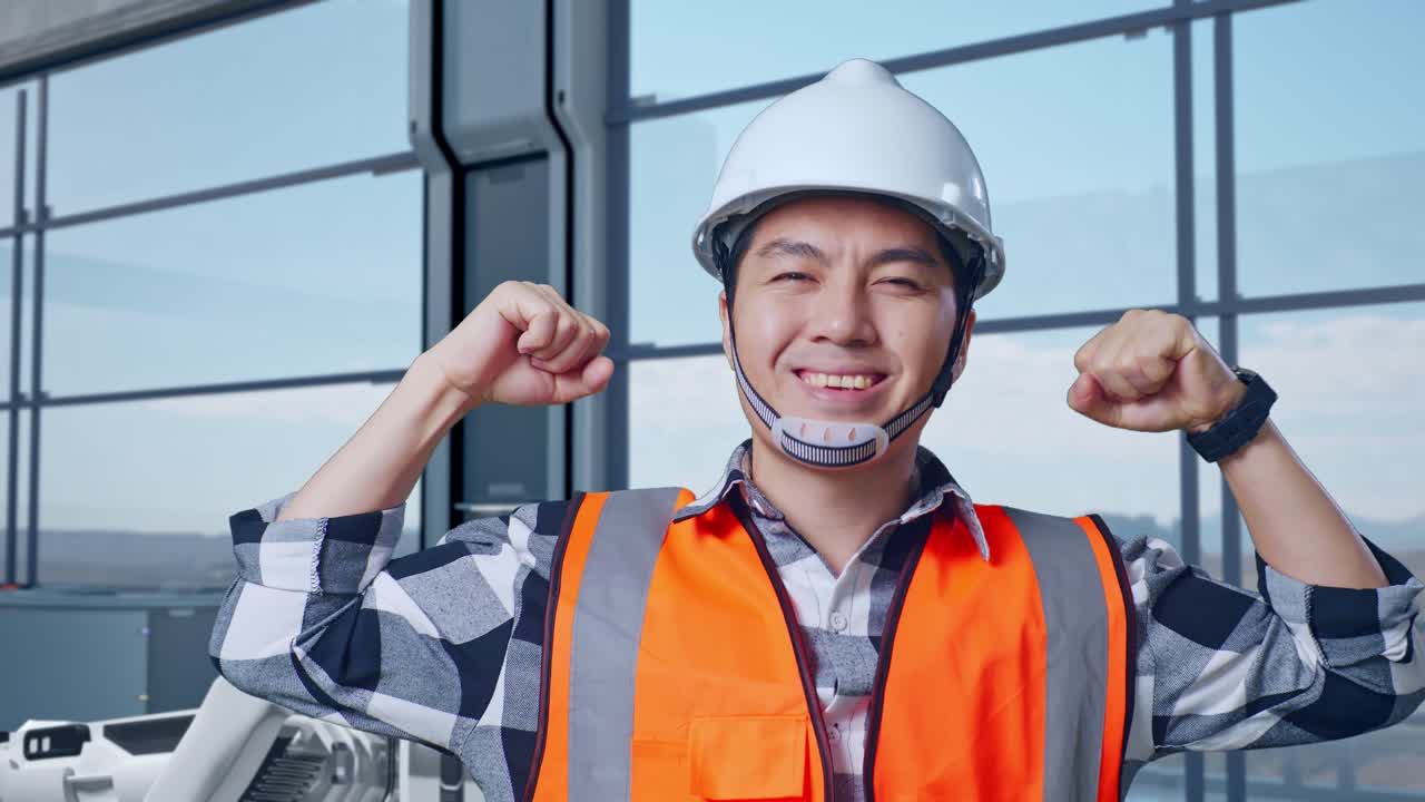 안전 헬을 착용한 아시아 남성 엔지니어가 하이테크 공장에서 로 기계 개념을 분석하며 이를 구부리고 카메라에 미소 짓고 있다.