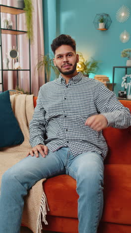 indian man smiling friendly at camera waving hands gesturing hello hi greeting at home on sofa