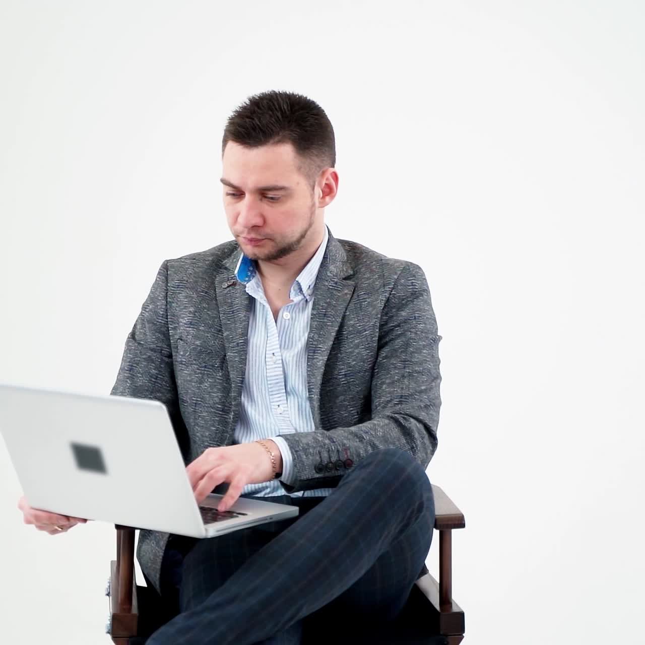 Young man sitting in a chair with laptop. Handsome businessman in stylish costume working on a modern computer, isolated in studio. Freelance work.