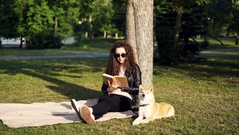 attractive girl student is reading book sitting on plaid under tree in city park with her puppy lying near and enjoying sunlight. hobby, leisure and animals concept.