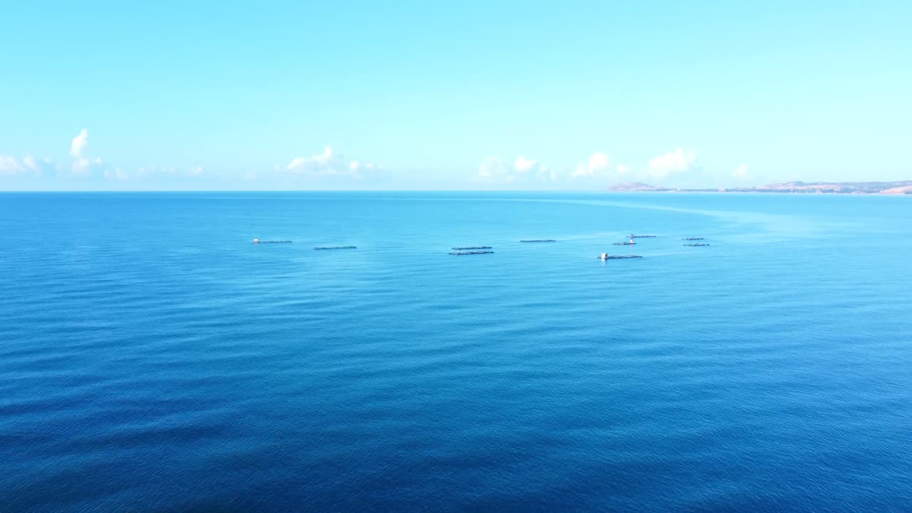 ベトナム南部フーコック島付近、穏やかな青い海の上空をドローンが空撮。遠くの海岸線と澄み切った空を背景に、養殖用の浮き漁業プラットフォームが映し出されている。