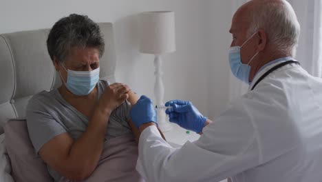médico caucásico de alto nivel vacunando a una mujer afroamericana de alto nivel en casa ambos con máscaras faciales