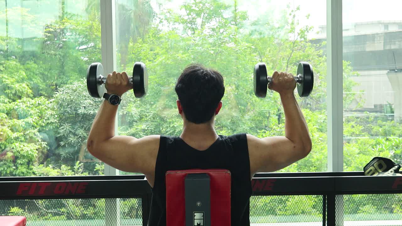 A man exercises with dumbbells in a gym, facing a large window with a green view
