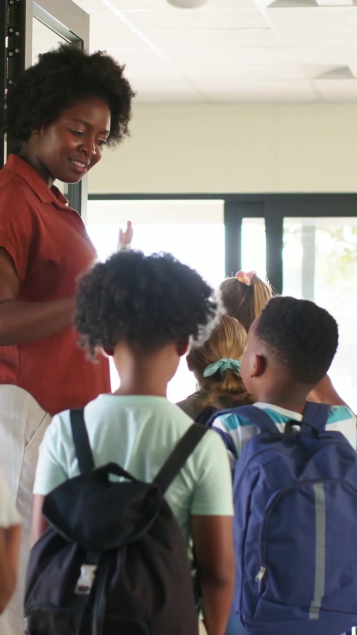Video verticale: Apre la porta, un'insegnante con una camicetta color ruggine guida bambini con zaini in classe