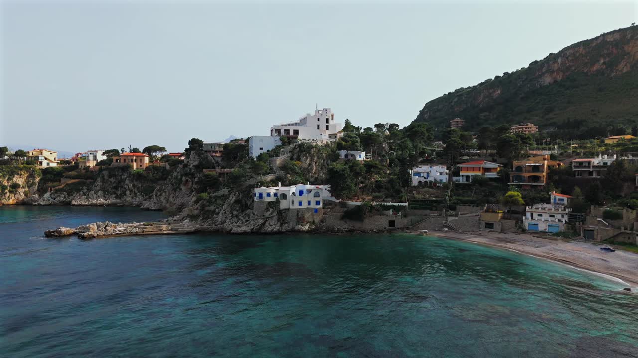 Drone shot of a seaside village near Porticello, Sicily, showing white and colorful houses, cliffs, and a beach with calm turquoise water under a clear sky
