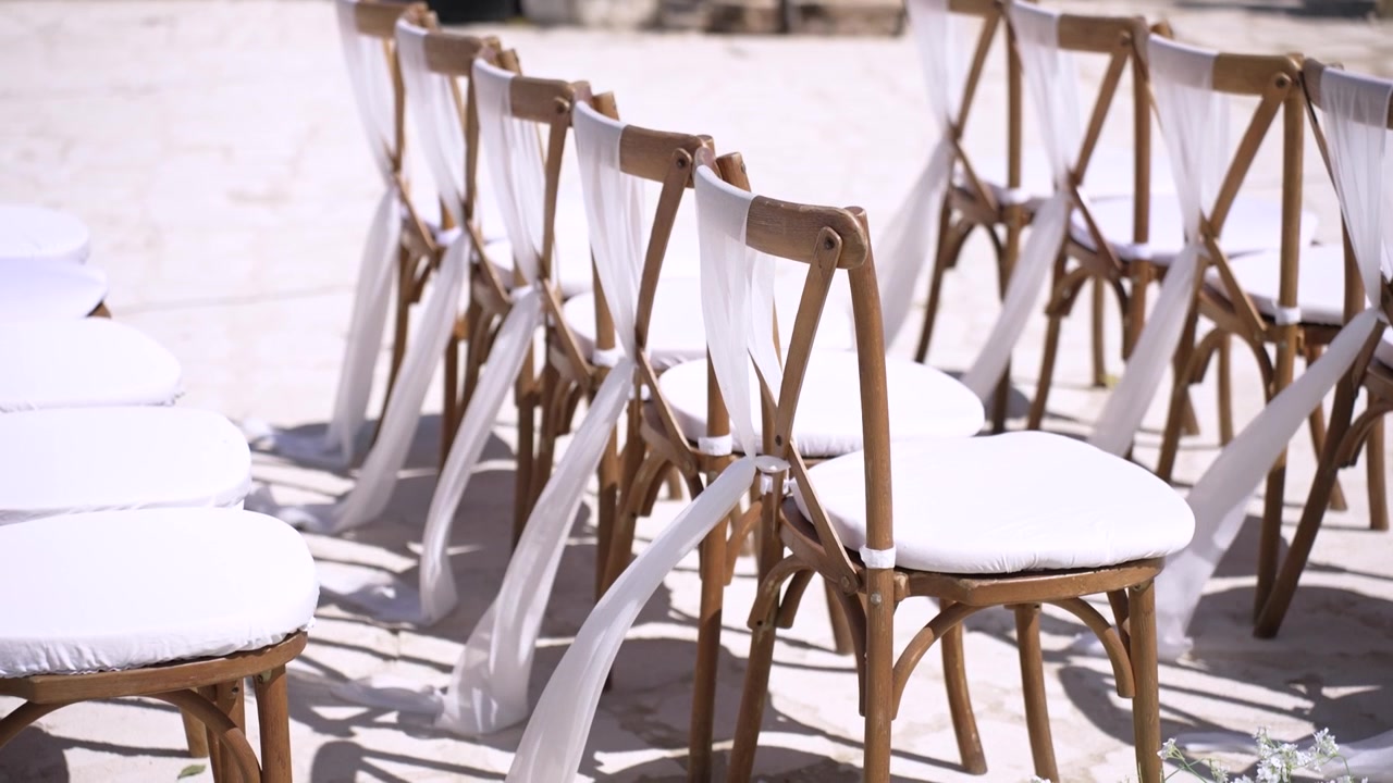 Des chaises sont disposées en extérieur pour une cérémonie de mariage. Chaque chaise est ornée d'un tissu blanc noué à son dossier. Le sol est pavé et prêt à accueillir les invités.