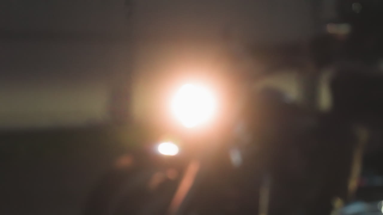 Close up of motorcycle headlight shining brightly as night riders move fast along road beside tall fence, blurred details highlighting speed and glowing light in dark evening environment