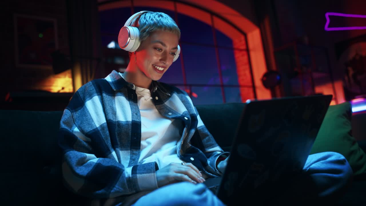 Young Beautiful Woman Wearing Headphones and Working from Home on Laptop Computer in Cozy Stylish Loft Apartment. Creative Female Smiling, Checking Social Media, Browsing Internet. Handheld Shot.