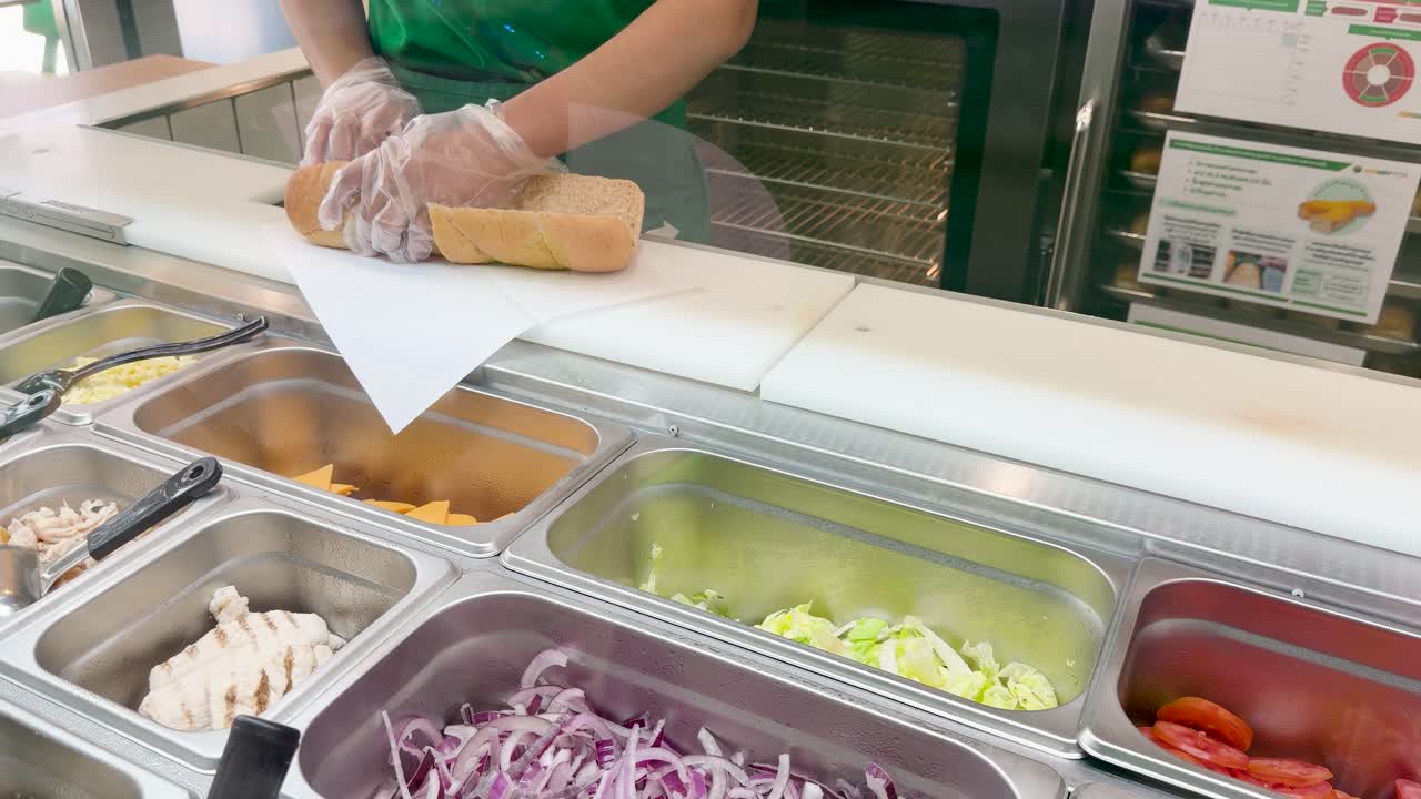 Un lavoratore che indossa guanti protettivi affetta un panino grigliato a un banco di sandwich commerciale.