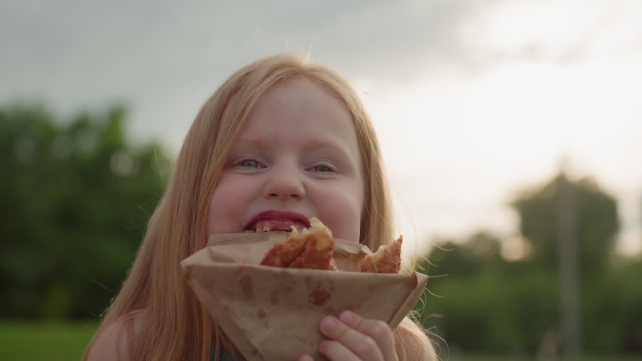 portrait of joyful kid holding pastry cutting paper wrap with teeth while chewing playfully on green outdoor background expression of fun and mischief during picnic moment