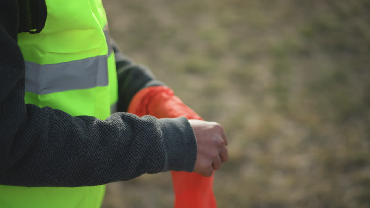 Worker in dark sweater and high visibility vest putting on bright orange protective gloves outdoors, preparing for safe work in construction with focus on hand movement and safety gear