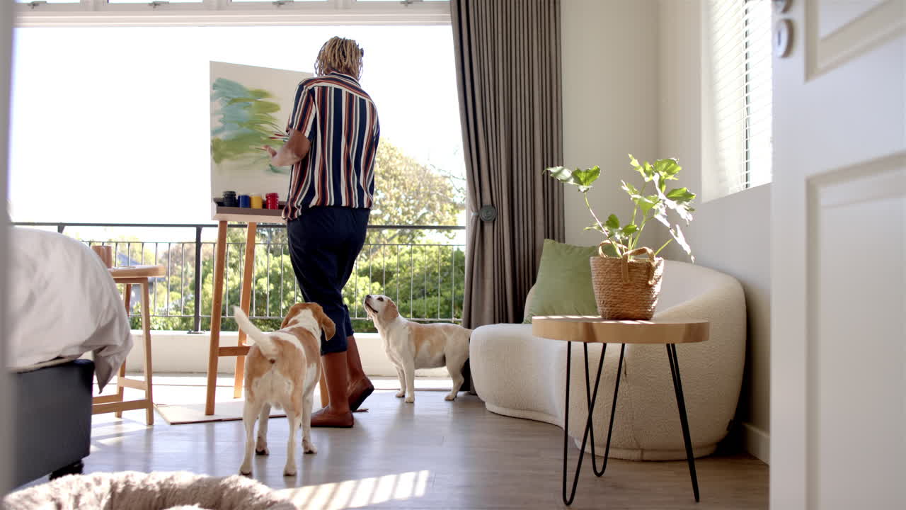A senior African American woman is painting on canvas, accompanied by two dogs