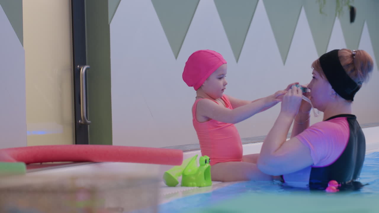 Swim instructor showing infant how to wear swimming goggles by pool, gentle guidance with hands, careful strap adjustment, bright light on water, warm family moment, supportive teaching scene
